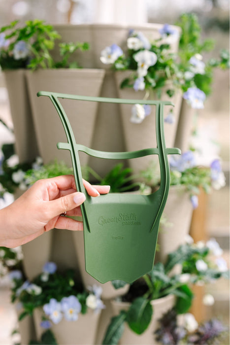 Hand holding a green GreenStalk garden trellis accessory in front of a vertical planter with blooming flowers and plants.