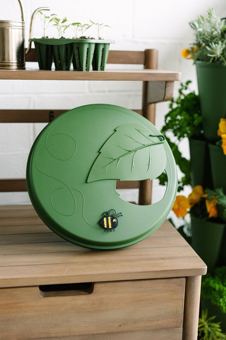 Green bee waterer with leaf design & bee accent on wood shelf, garden seedlings, watering can, and potted plants.