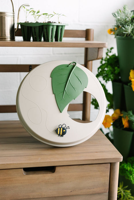 Decorative garden plate with green leaf, bee, and cutout on wood table. Seedlings, watering can, potted herbs.