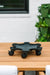 Black wheeled plant caddy on a wooden patio table. Vertical garden with green pots and white flowers in background.