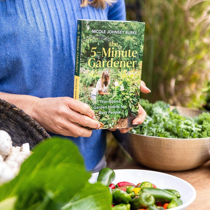 "The 5-Minute Gardener" book by Nicole Johnsey Burke, on year-round garden habits, held near fresh kale, peppers, and mushrooms.