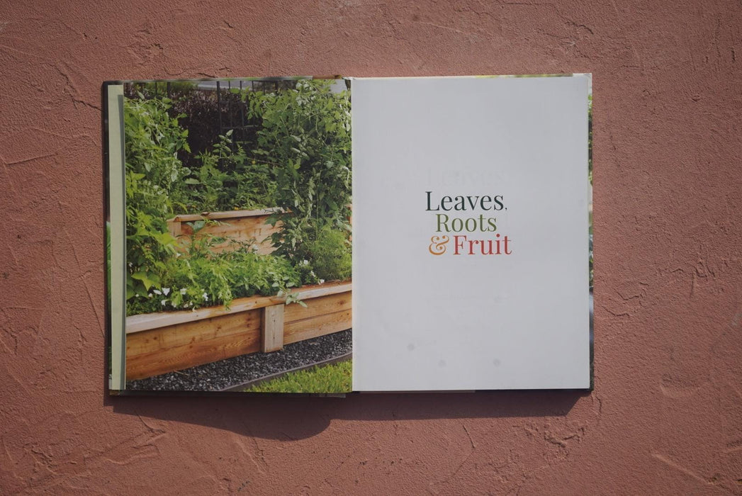 Open gardening book "Leaves, Roots & Fruit" showing a vibrant raised garden bed with lush edible plants.