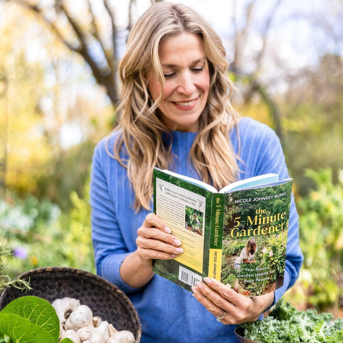 Happy woman reading "The 5-Minute Gardener" by Nicole Johnsey Burke in a vibrant garden with fresh garlic and kale.