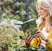 Blonde woman reads "The 5-Minute Gardener" book by Nicole Johnsey Burke in a garden, holding fresh sage plants. Easy garden tips.