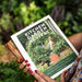 Hands holding "Kitchen Garden Revival" book, showing a woman in a lush edible garden. Guide for small-scale, low-maintenance gardening.