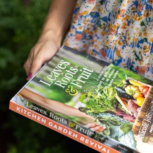 Hand holding "Leaves, Roots & Fruit" kitchen garden book. Cover shows fresh organic vegetables & greens.