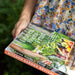 Hand holding "Leaves, Roots & Fruit" kitchen garden book. Cover shows fresh organic vegetables & greens.