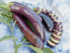 Long purple Asian eggplants, whole and sliced, with fresh green leaves on a blue patterned cloth.