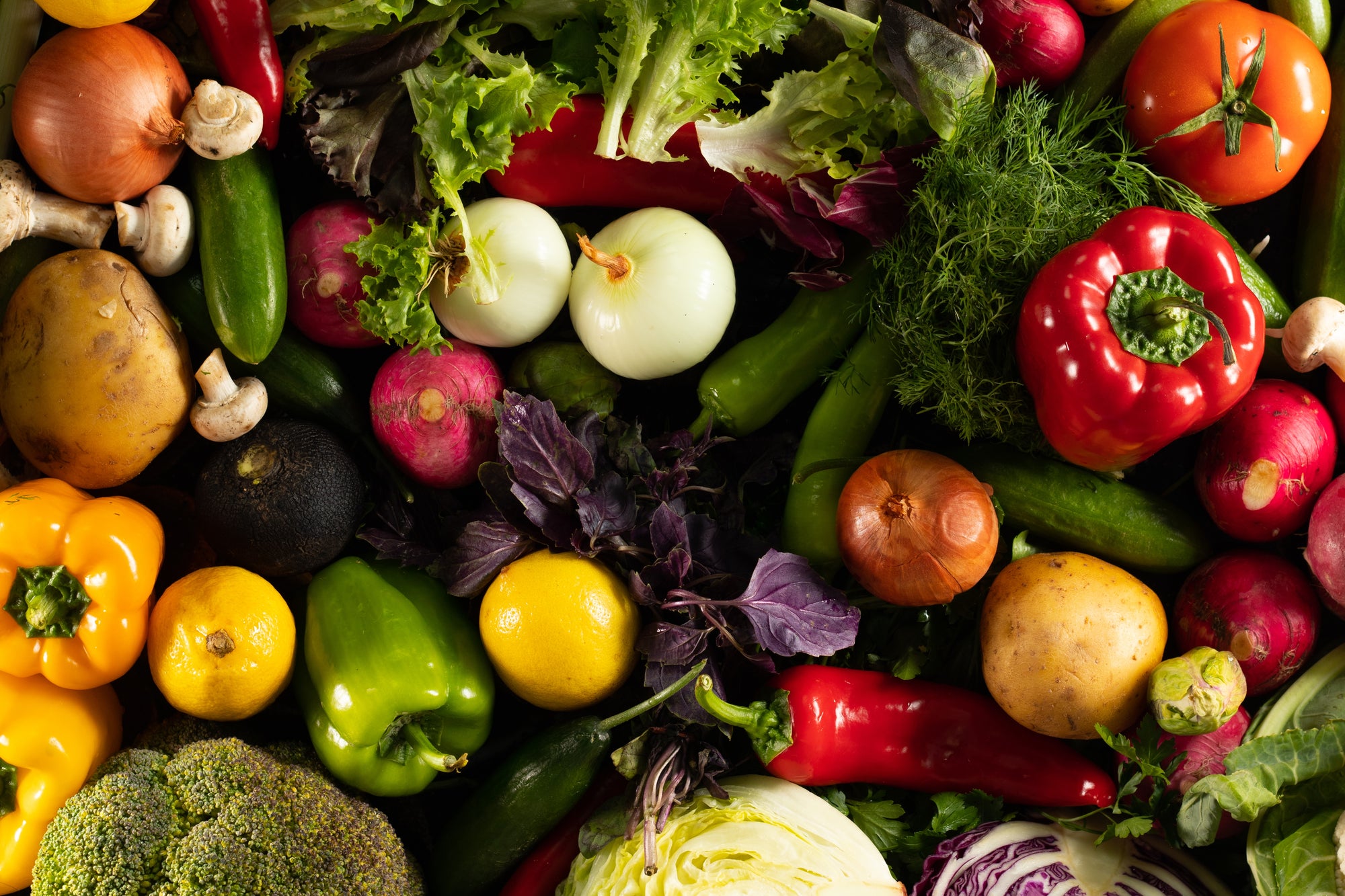 Vibrant mix of fresh organic produce: bell peppers, onions, tomatoes, lettuce, broccoli, mushrooms, potatoes, radishes, lemons, herbs.