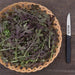 Fresh green and purple baby mustard greens in a woven basket, next to a serrated knife on a wood table.