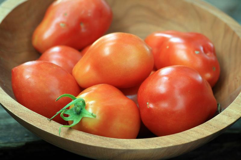 Ripe red and orange plum tomatoes with green stems in a natural wooden bowl. Fresh garden produce.