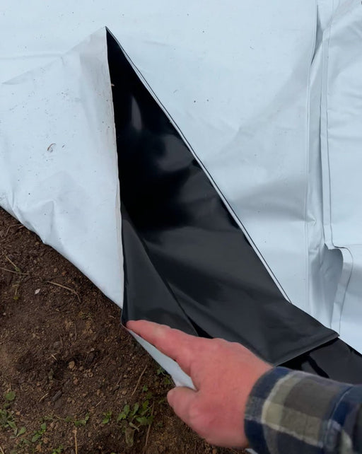 Hand lifting white agricultural mulch film, revealing black layer on garden soil.