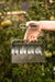 Gardener's hand holding a shiny metal multi-seed planter tool in a vibrant garden with green tomatoes. Efficient planting.