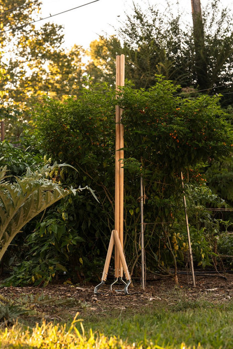 Tall wooden garden cultivator with three metal weeding hoes stands in a sunny vegetable garden among lush plants.