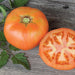Large peach-orange heirloom tomato, whole and sliced, showing juicy interior on rustic wood with green leaves.