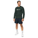 Male model in dark green Seedtime long-sleeve shirt. Comfortable men's athletic wear, navy shorts, white sneakers.