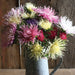 Vibrant spiky aster bouquet: pink, yellow, red, white, purple garden flowers in a rustic metal pitcher on wood.
