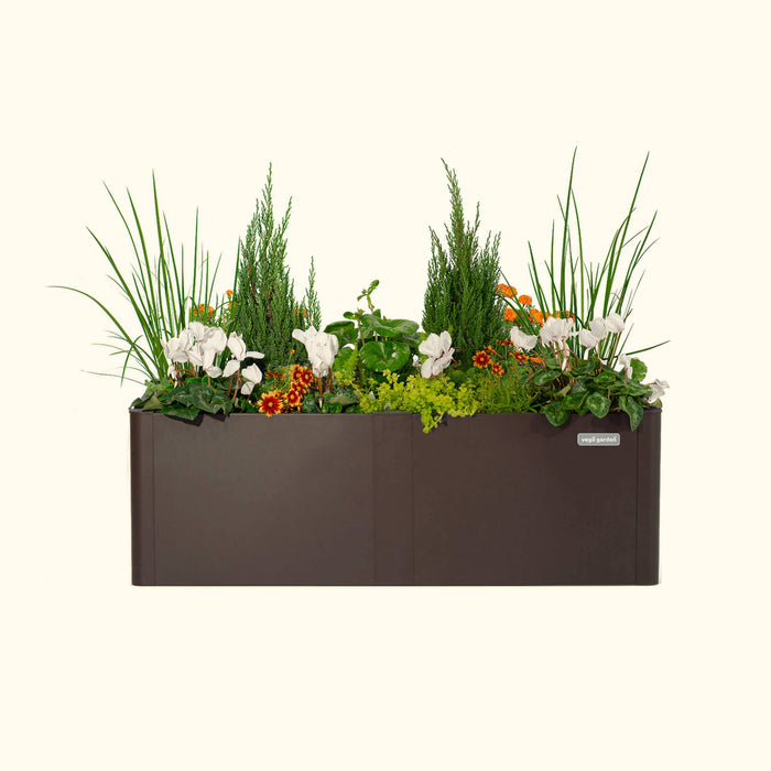 Vego Garden brown planter box with lush green grass, evergreen shrubs, white cyclamen, and vibrant orange flowers.