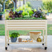 Cream raised garden bed on casters with leafy greens, pink flowers, gardening tools, and brass watering can.