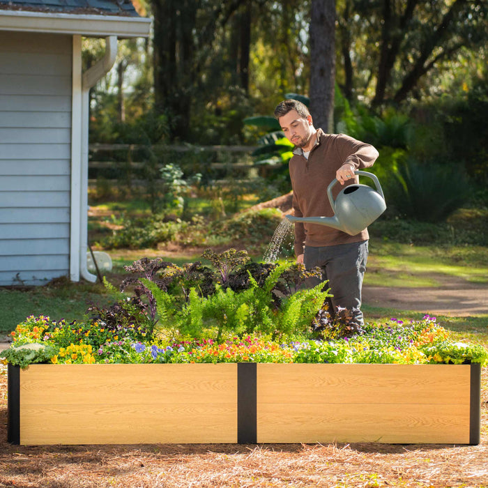 Man watering vibrant flowers and plants in a modern wood-look raised garden bed, ideal for backyard gardening.
