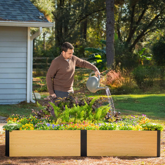 Man watering a composite raised garden bed filled with colorful flowers and leafy greens in a sunny backyard garden.