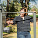 Woman installing grey metal garden trellis panel. Sunny backyard, greenhouse, and raised bed in background.