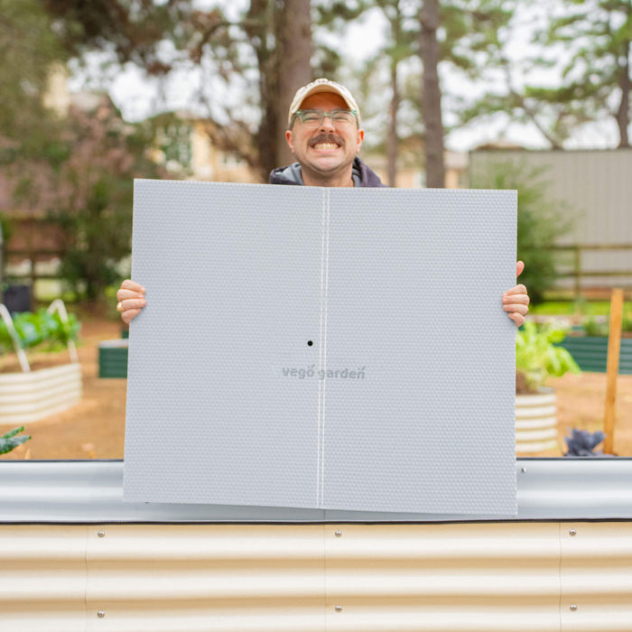 Smiling man holds Vego Garden textured panel for a beige corrugated metal raised garden bed in a backyard.