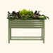 Sage green elevated metal garden bed with storage shelf, filled with kale, purple leafy greens, ferns, and pink flowers.
