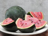 Fresh whole dark green watermelon with several pink flesh slices and visible seeds on a white plate.