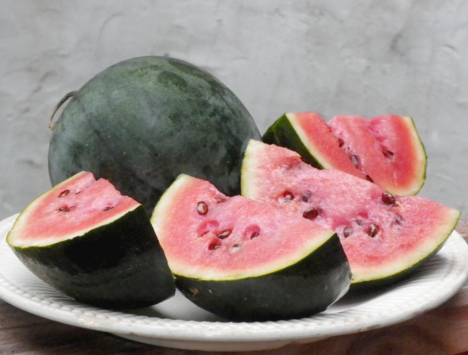 Fresh whole dark green watermelon with several pink flesh slices and visible seeds on a white plate.