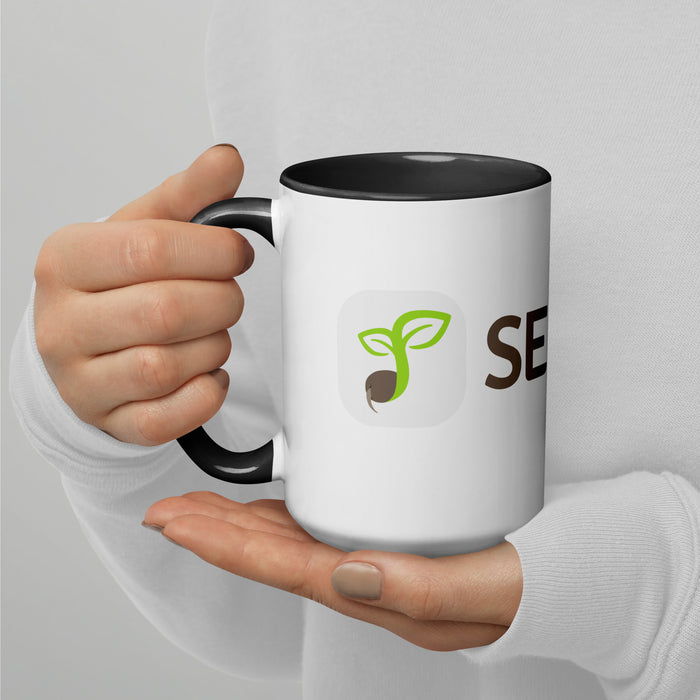 Person holding white two-tone mug with black handle, green sprouting seed logo, and 'SEED' text.