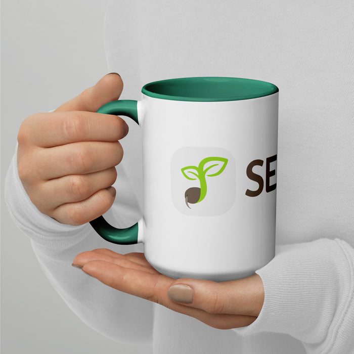 White ceramic mug with green handle, sprout seed logo, and partial "SEED" text, held by hands. Great for coffee or tea.