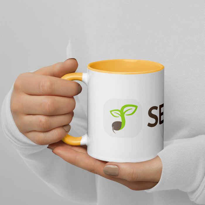 Hands hold white coffee mug with yellow handle, green seed sprout logo, and "SEED" text. Eco-friendly drinkware.
