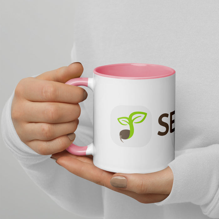 Hands holding a white and pink coffee mug with a green sprouting seed logo and 'SEED' text. Eco-friendly drinkware.