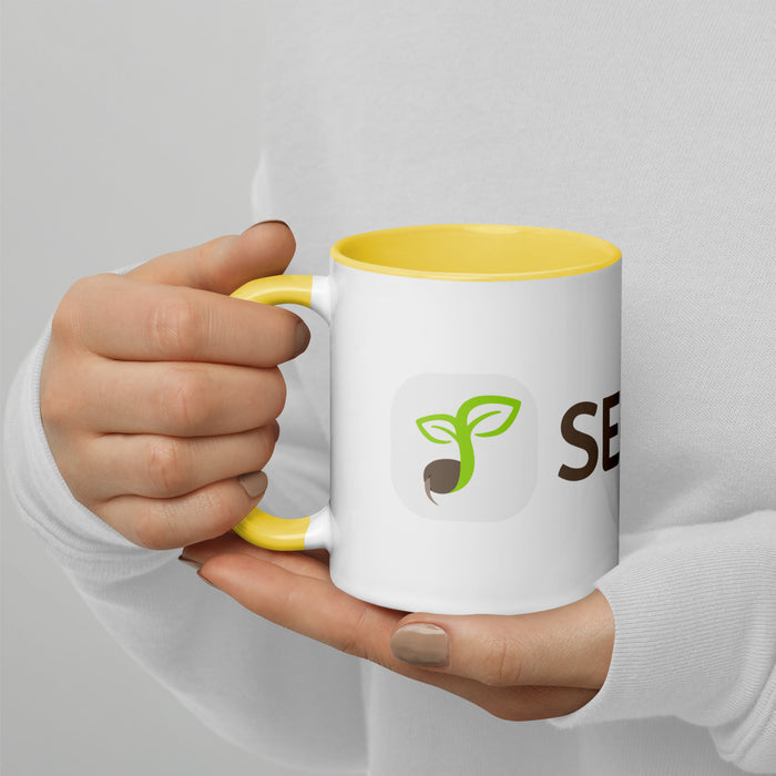 White mug with yellow handle, green seed sprout logo, and 'SE' text, held by hands. Branded coffee cup.