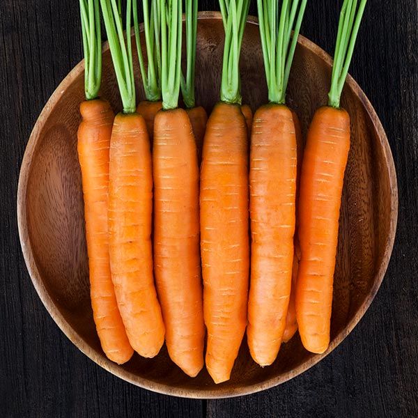 Eight fresh, vibrant orange carrots with green tops in a natural wooden bowl on a dark wood surface.