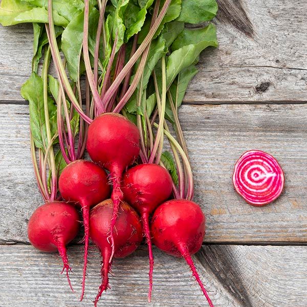 Fresh red beets with green tops and roots, one sliced Chioggia beet showing red and white stripes, on rustic wood.