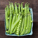 Fresh green pea pods, one open with bright peas, in a market carton on dark wood. Raw garden vegetables.