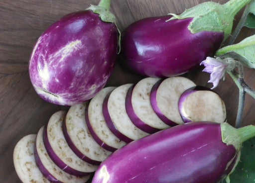 Varied purple eggplants: one striped, two solid, plus fresh slices, flower, and leaves on a wooden surface.