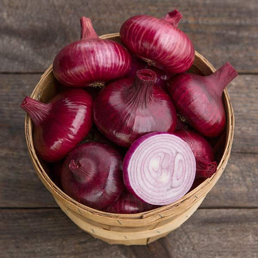 Fresh red onions in a wooden basket on a dark wood surface, with one sliced purple onion showing its rings.